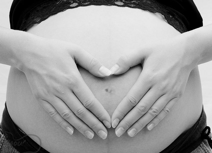 A black and white photo shows a pregnant woman forming a heart shape with her hands around her belly